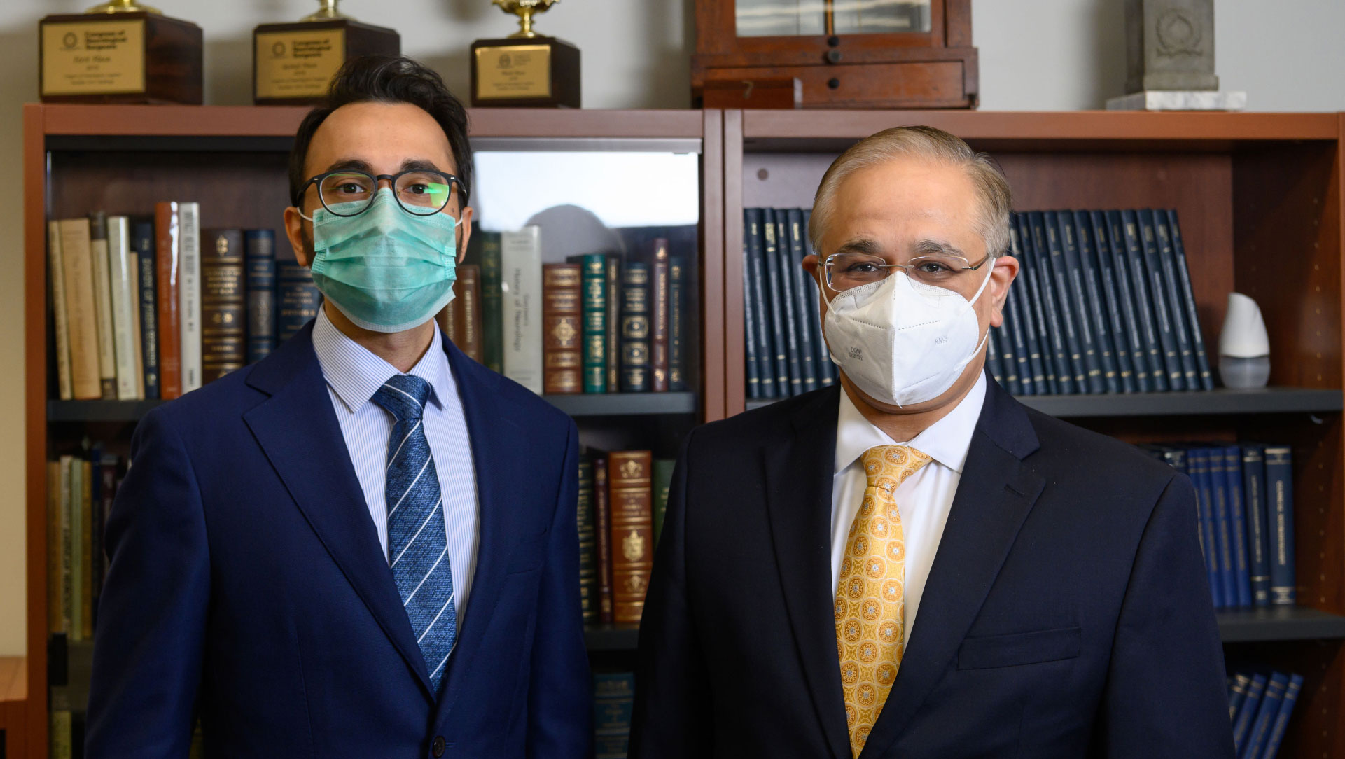 Dr. Farhan Mirza (left), a younger man with dark hair and a beard with glasses, and Dr. Siddharth Kapoor (right), a middle aged man with glasses, both dressed in suit jackets and ties with face masks on standing side by side in front of a bookshelf.