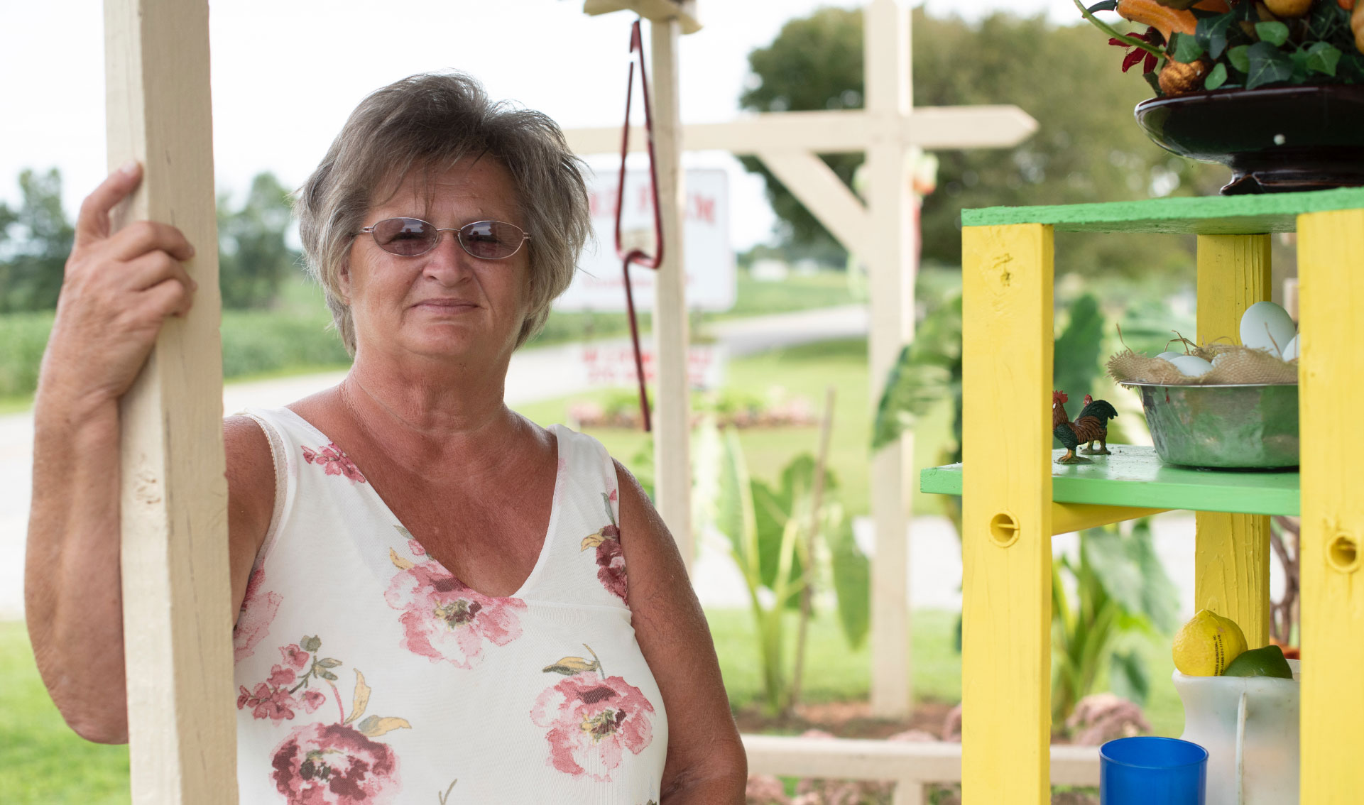 A portrait photo of Charlotte, dressed in a white tank top with pink flowers, standing on her porch.