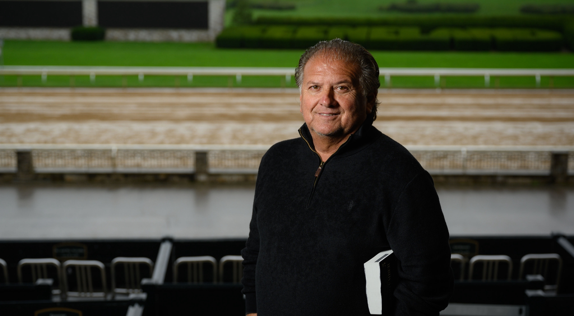 Omar Trevino, an older Hispanic man with combed back gray hair, smiles for the camera while standing in front of the racetrack. He is wearing a long-sleeve black pullover sweater.