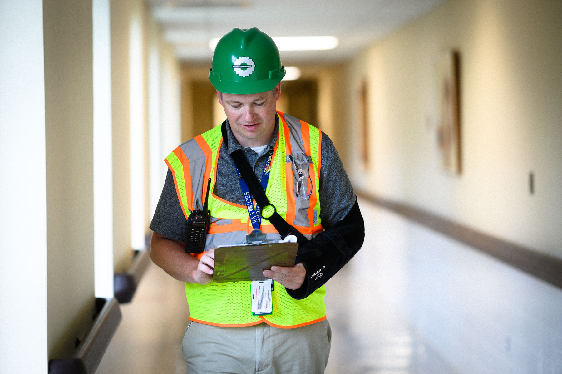 Leonard walks down a hall with a clipboard while wearing a safety vest and construction helmet.